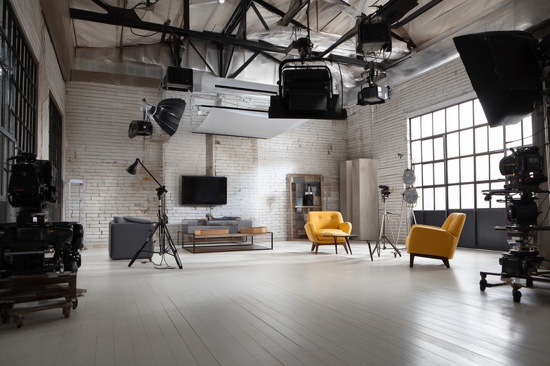 Bright industrial loft studio interior for photo and video production featuring professional camera equipment, studio lights, large windows, exposed brick walls and yellow armchairs.