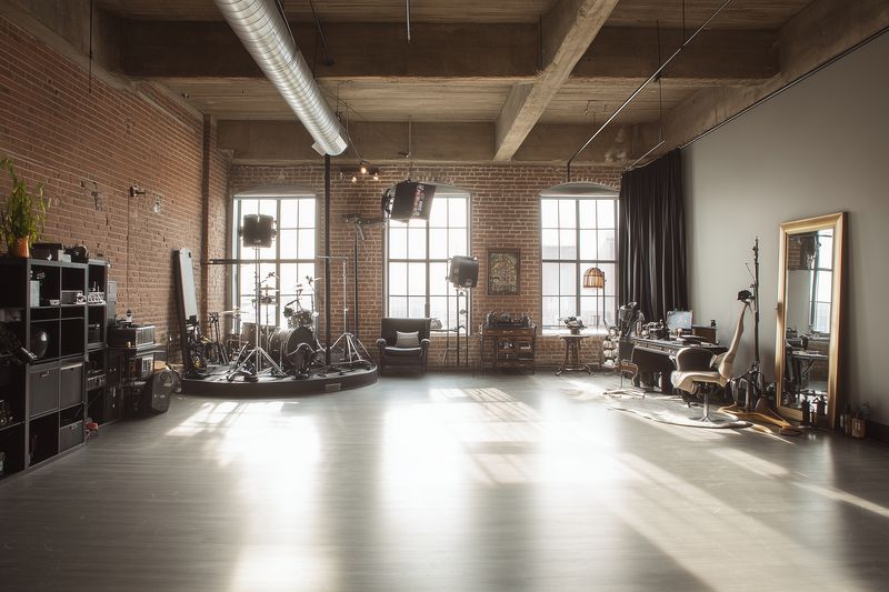 Bright industrial loft photography studio with exposed brick, large windows and professional lighting equipment arranged around vintage furniture and open hardwood floor.