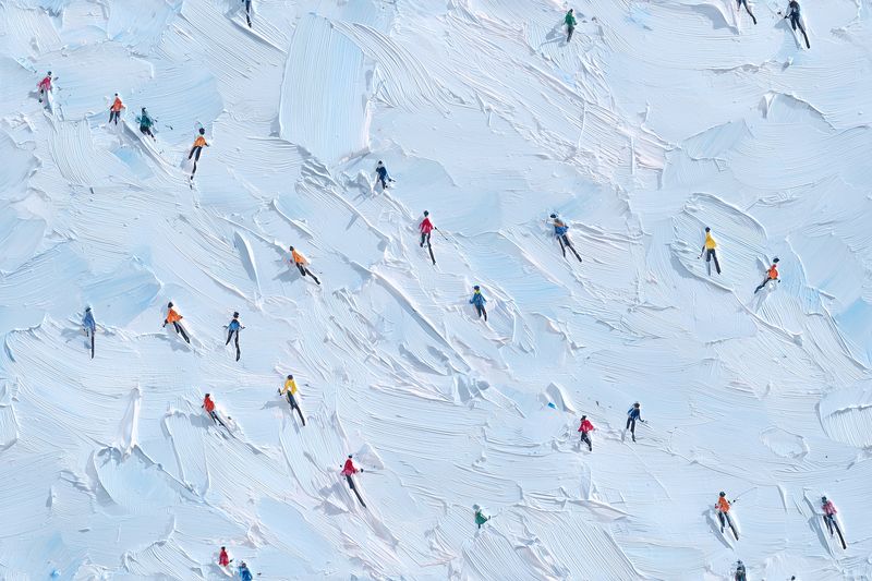 Aerial view of numerous skiers descending a snowy slope, leaving crisscrossing tracks across pristine powder while colorful jackets punctuate the bright winter landscape.