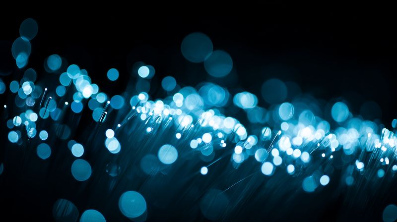 Close-up abstract photograph of glowing blue bokeh lights resembling fiber optic strands, creating a soft luminous texture and sparkling highlights against a deep dark background for creative use.