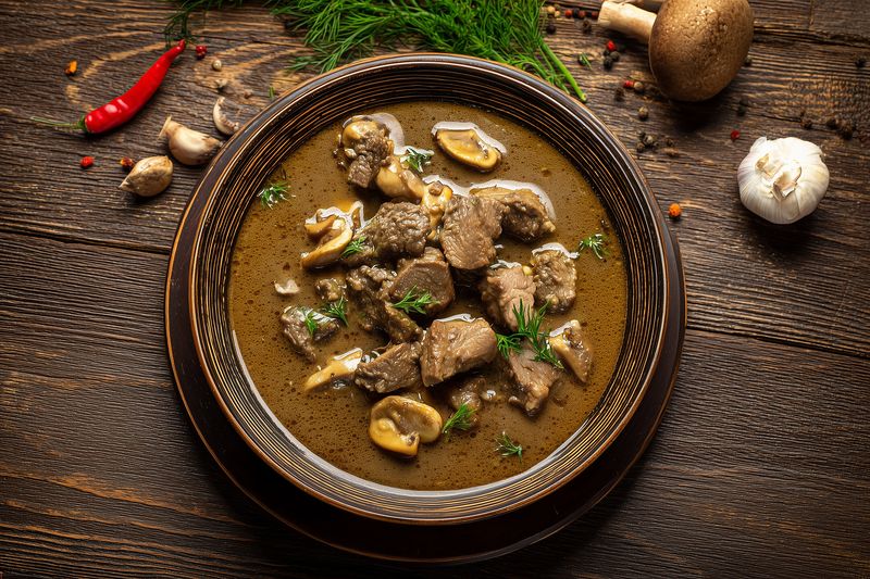 Hearty beef and mushroom stew served in a rustic bowl on a wooden table, garnished with fresh dill and garlic cloves, warm savory broth perfect for cozy comfort meals.