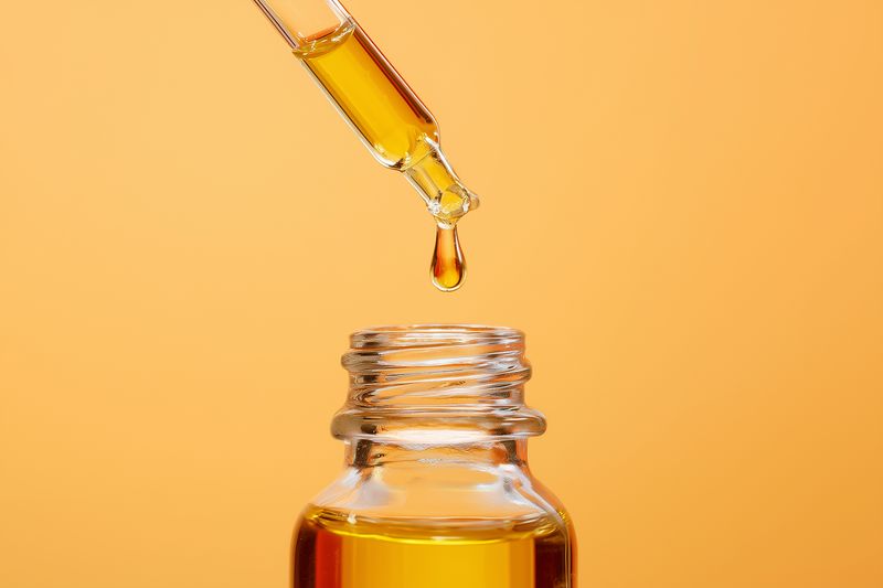 Close-up of a glass dropper releasing a golden oil drop into a small clear bottle against a warm orange background, highlighting texture, clarity, and graceful liquid motion.