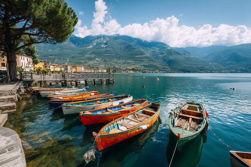 Colorful wooden boats moored along a tranquil lakeside with clear turquoise water, sunlit promenade and distant mountain range under a bright sky with fluffy clouds.