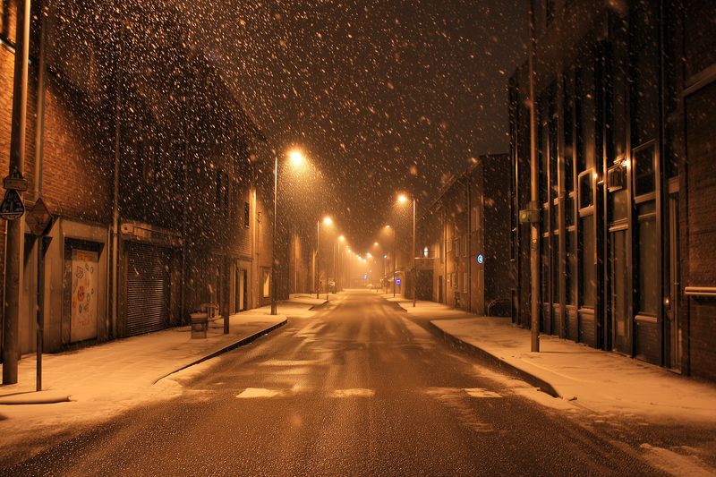 Empty urban street at night during a gentle snowfall, warm streetlights casting golden reflections on wet asphalt, a quiet moody scene with falling snowflakes and misty glow.
