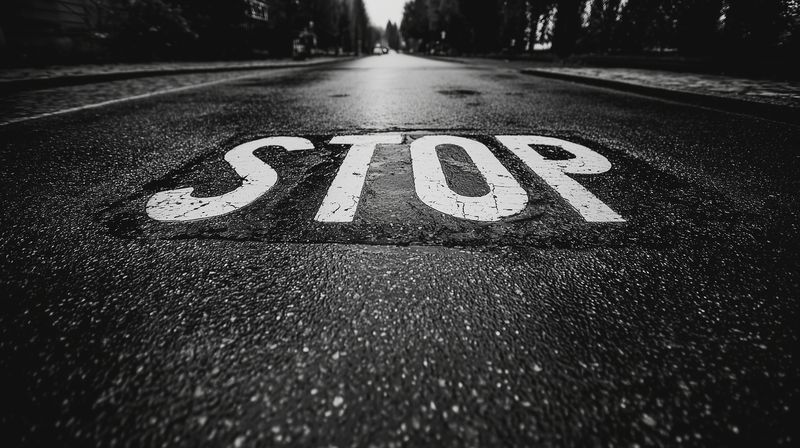 Low angle black and white photo of a wet asphalt road with a large painted STOP marking in the foreground, textured surface and dramatic vanishing perspective.