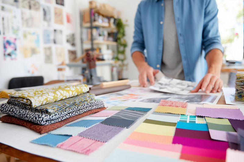 Textile designer arranging colorful fabric swatches and paper color samples on a bright studio table, planning patterns and layouts with assorted samples and creative tools.