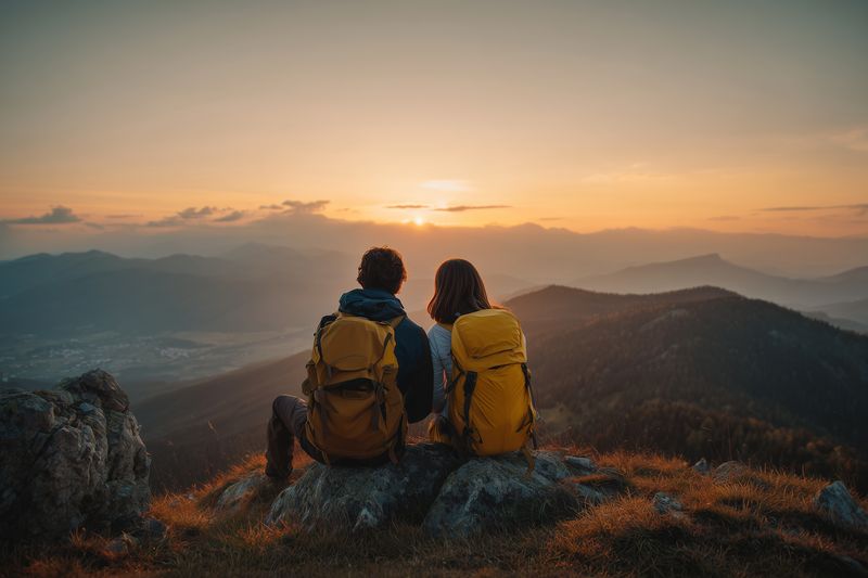 Two hikers with backpacks sit on a rocky ridge watching a golden sunset over layered mountain ranges, sharing a peaceful moment of adventure and natural beauty together.