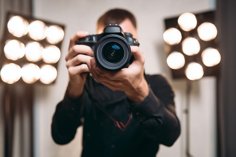 Portrait of a photographer holding a DSLR camera toward the viewer in a studio environment with blurred background lights, emphasizing the camera lens and hands.