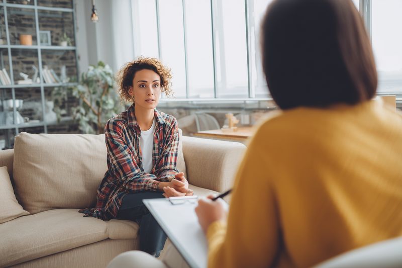Young woman seated on a sofa during a counseling session, speaking with a therapist in a cozy modern office, expressing emotions and seeking understanding and emotional support.