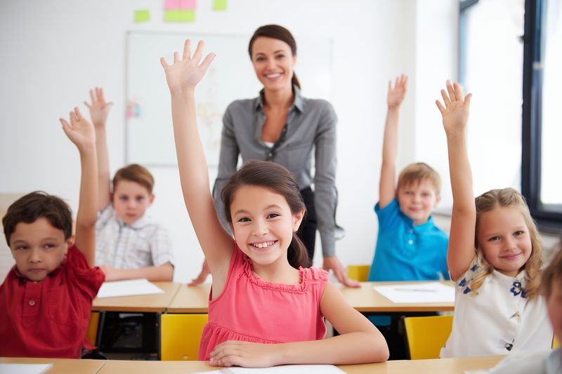 A group of happy elementary students raise their hands eagerly in a bright classroom while a smiling teacher stands at the front, encouraging participation and active learning.