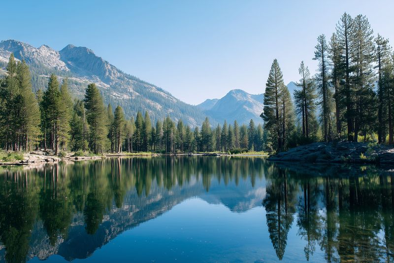 Calm mountain lake surrounded by pine forest and rugged peaks, reflecting clear blue sky and evergreen trees in pristine water, creating a peaceful alpine landscape.