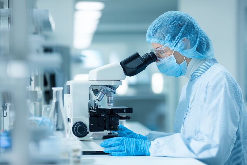 Laboratory scientist in protective gear examines slides under a microscope in a sterile clinical lab, performing precise biomedical research and diagnostic testing.