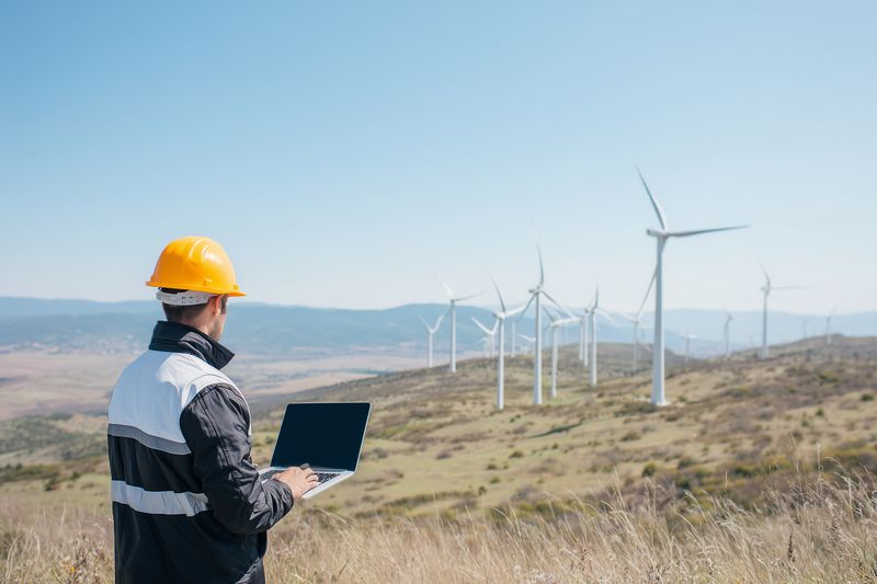 Technician in a safety helmet using a laptop to monitor wind turbines on a grassy hillside, renewable energy inspection, field maintenance and remote performance analysis under clear sky.