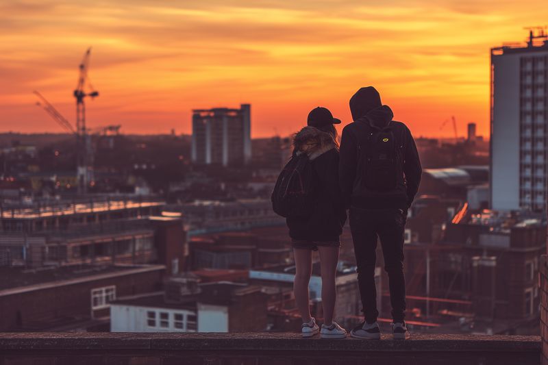 Silhouetted couple stands on a rooftop overlooking an urban skyline at golden sunset, warm light casting long shadows as city rooftops glow in orange and red hues, backpacks visible.