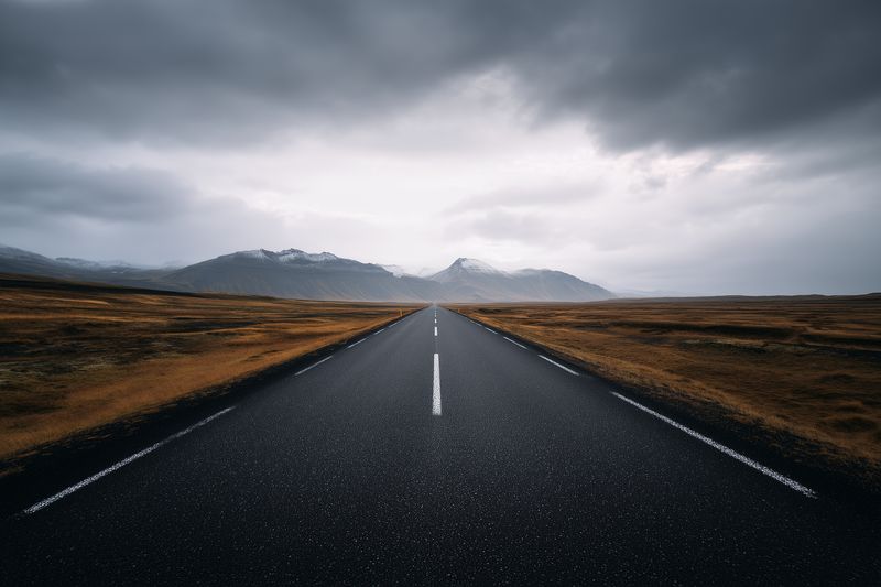 A lonely asphalt road stretches toward distant snow-capped mountains under a dramatic moody sky, flanked by barren golden fields and rugged plains, conveying solitude and wanderlust.