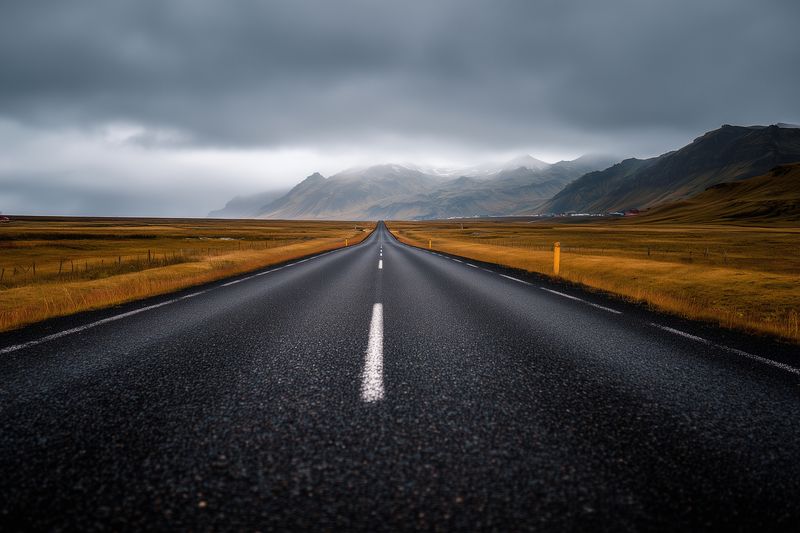 Dramatic empty asphalt road stretching toward distant misty mountains under heavy storm clouds, framed by golden grassy plains and moody atmospheric light in a remote landscape.
