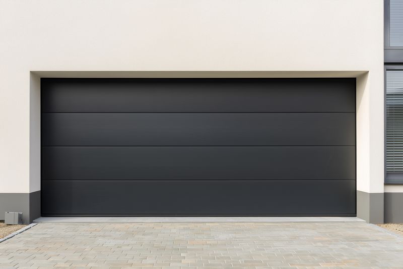 Contemporary closed dark garage door set into a clean minimalist facade of a residential building, featuring horizontal panels, sleek matte texture, subtle symmetry and a paved concrete driveway.
