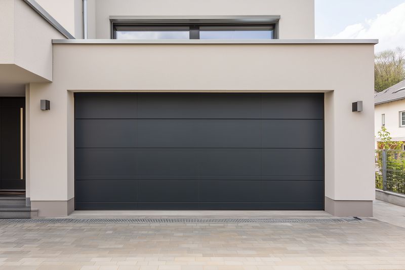 Modern residential exterior featuring a closed dark garage door on a contemporary house facade, smooth stucco walls, minimalist design and clean driveway in daylight.