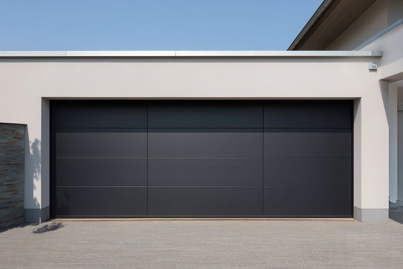 Modern residential garage with closed dark sectional door set in a minimalist facade, clean driveway and subtle architectural details evoking contemporary suburban home exterior.