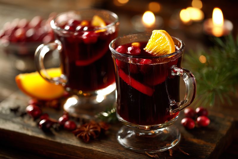 Warm mulled wine in clear glass mugs garnished with orange slices, cranberries and aromatic spices on a rustic wooden table lit by soft candlelight, evoking a cozy festive mood.