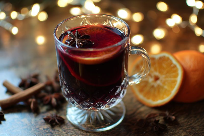 Warm spiced mulled wine in a glass mug with star anise and citrus slice, cozy holiday beverage set on rustic table with bokeh lights and cinnamon sticks for festive atmosphere.