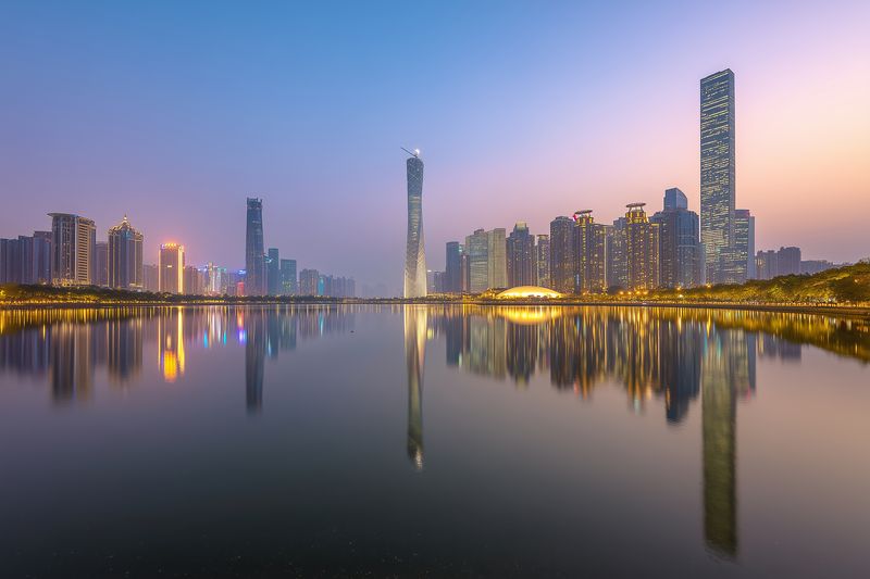 Panoramic twilight city skyline reflected on serene water, modern highrise towers and illuminated windows glow against a soft pastel evening sky in a peaceful urban waterfront scene.