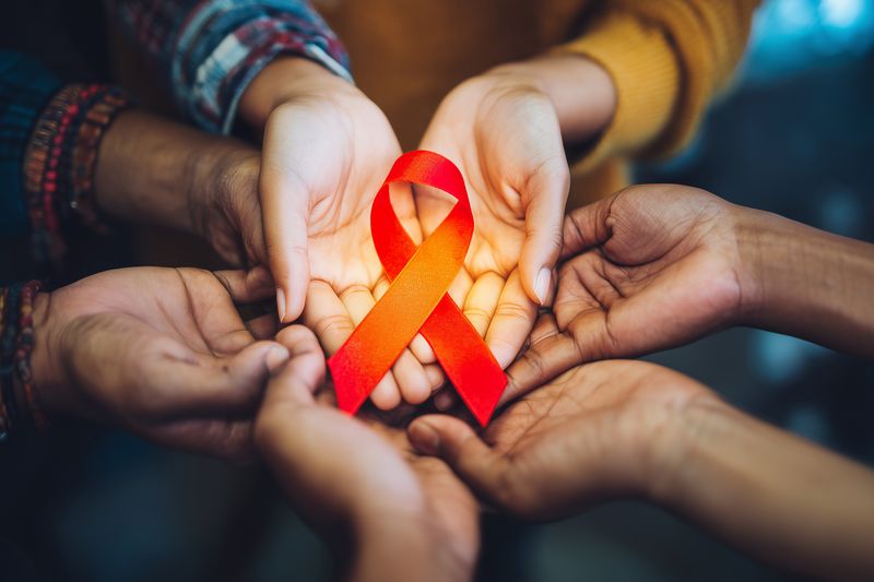 Closeup of diverse hands gently holding a red awareness ribbon, symbolizing compassion, support and solidarity for HIV and AIDS prevention, care, health education and hope.