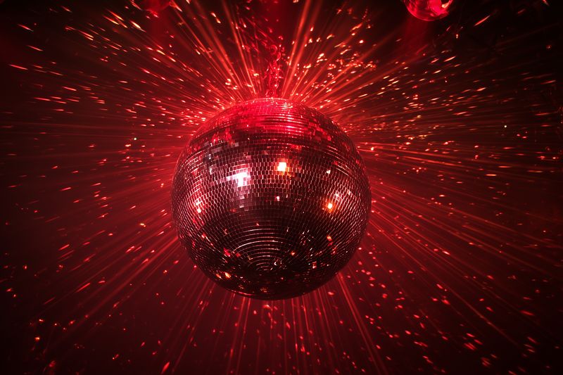 Mirrored disco ball bathed in red light, throwing countless sparkling reflections and radial beams across a dark ceiling, evoking lively dance club energy and festive celebration.