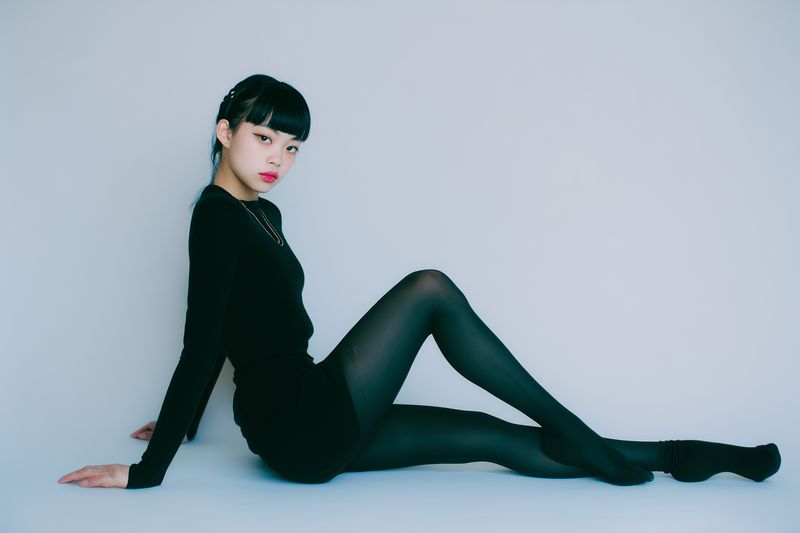 Studio portrait of a female model in a black dress and sheer stockings seated against a clean white background, striking an elegant minimalist pose with a poised expression.