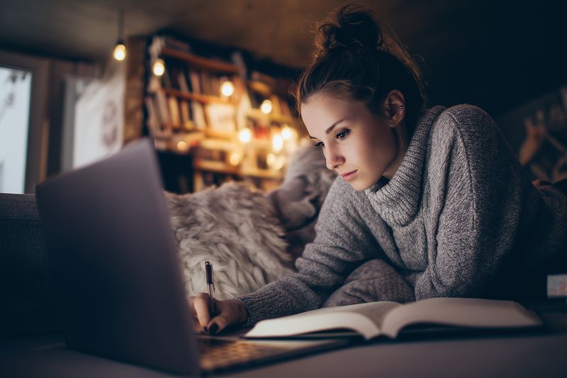Young woman studying late at night on a cozy sofa with laptop and notebook, focused expression, warm ambient lighting and soft background bokeh creating an intimate home workspace.