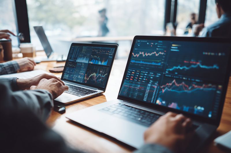 Two professionals analyze financial charts and market data on laptop screens in a modern café workspace, collaborating on trading strategies and investment analytics.
