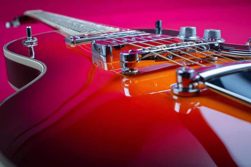 Close-up of a glossy red electric guitar showcasing chrome bridge, strings, pickups and control knobs, captured with dramatic studio lighting and a shallow depth of field.