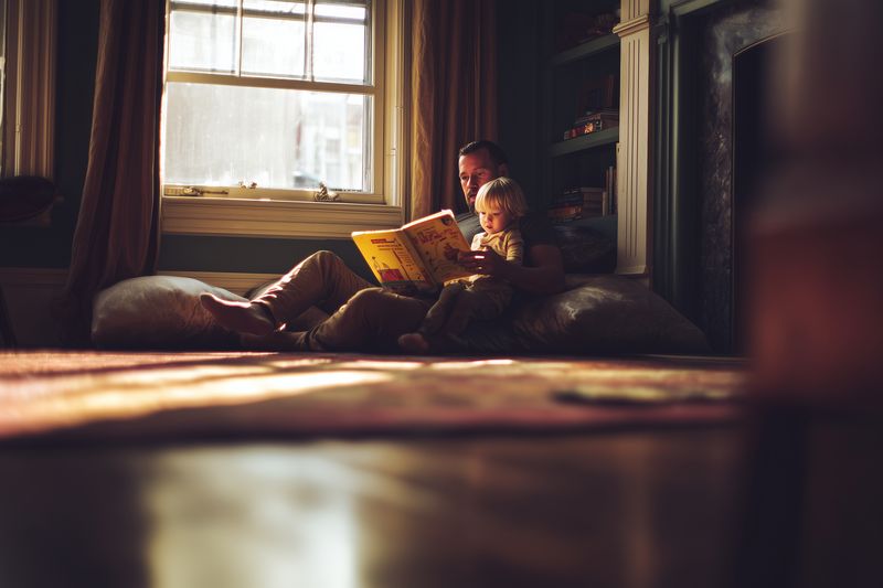 A parent and young child sit on cushions by a window in a warm living room, sharing a book while sunlight filters in, creating a cozy intimate family reading moment.