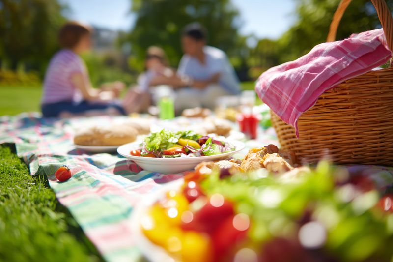Family enjoying a sunny outdoor picnic on a colorful blanket with fresh salad, fruit, bread and a woven basket, capturing a relaxed warm afternoon in a green park.
