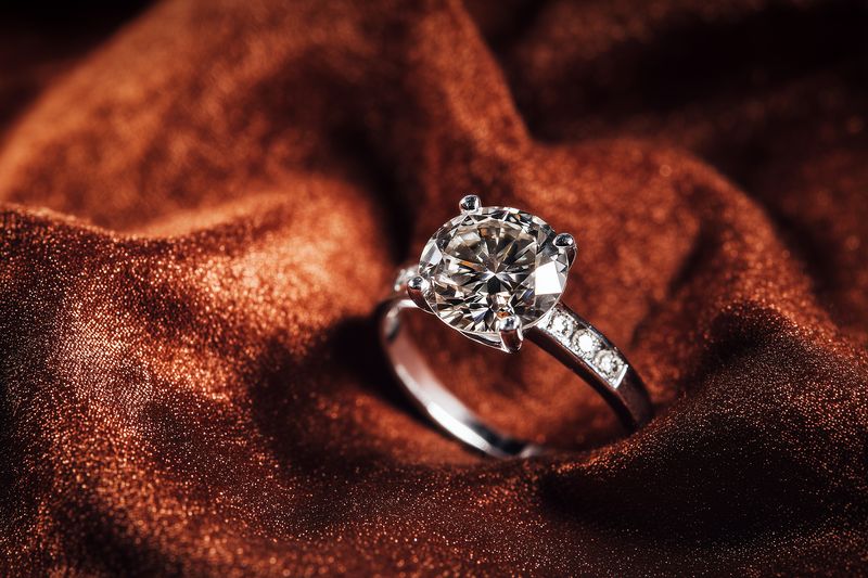 Close-up of a sparkling solitaire diamond engagement ring resting on luxurious brown satin fabric with glitter highlights, showcasing elegance, romance and fine jewelry detail.