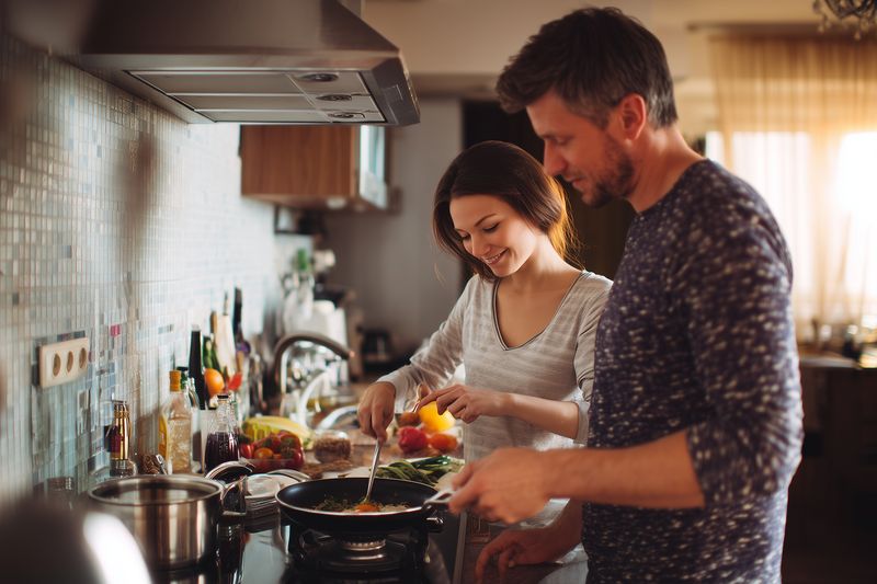 Young couple cooking together in a warm home kitchen, smiling as they prepare a meal, chopping vegetables and stirring a pan near a sunlit window, sharing a cozy domestic moment.