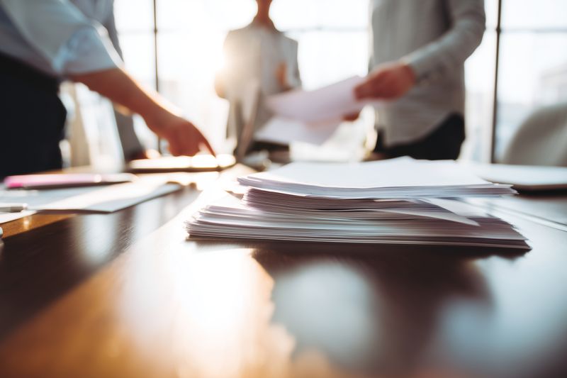 Close-up view of stacked documents and papers on a conference table as business colleagues discuss reports and exchange printed materials, warm sunlight and shallow depth of field suggest focus.