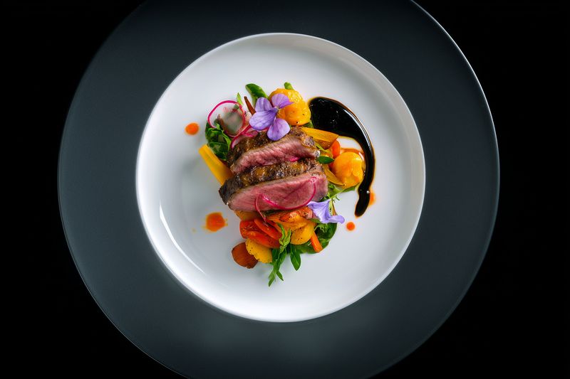 Top view of a gourmet plated beef steak with colorful vegetables, edible flowers and sauce on a white plate set on a dark textured charger for elegant restaurant presentation.