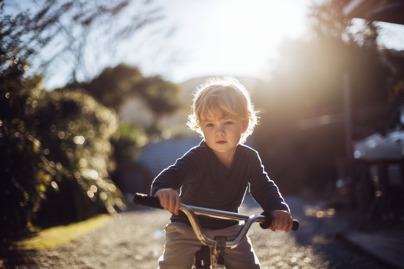 A young boy rides a small bicycle along a sunlit garden path, golden backlight illuminating his blond hair as he concentrates on balance and discovery in warm afternoon light.