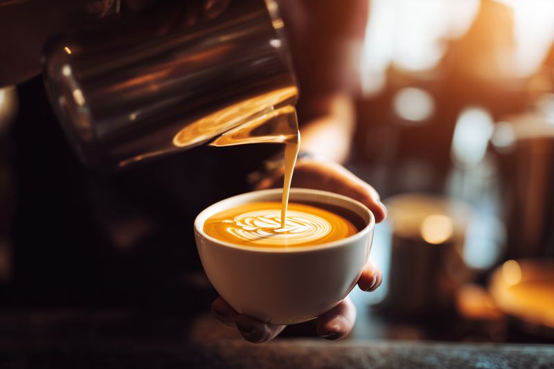 Barista pouring steamed milk to create latte art in a ceramic cup, warm cafe atmosphere with bokeh lights and rich crema, close-up of hands and coffee preparation.