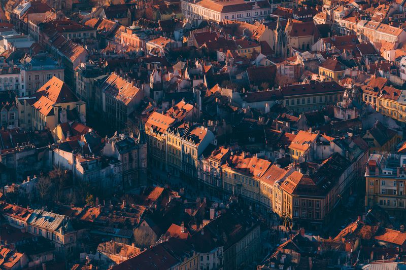 Aerial view of a dense historic cityscape with warm sunlight casting long shadows over terracotta rooftops, narrow streets and ornate facades forming a textured urban mosaic.
