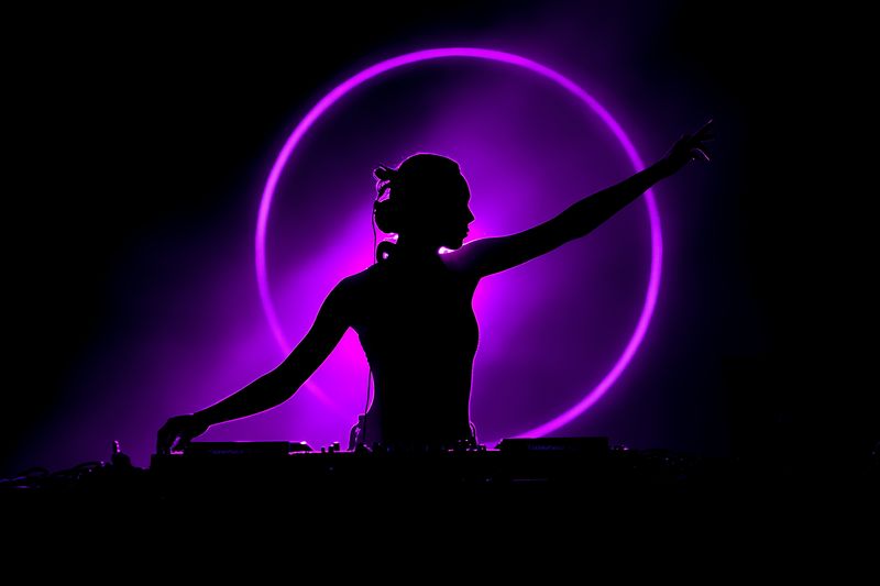 Silhouette of a female DJ performing at a nightclub, backlit by a glowing purple neon circle that creates dramatic contrast and an energetic atmosphere for a live music event.