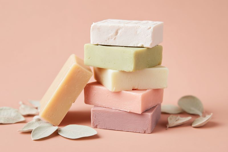 Stack of handmade pastel soap bars displayed on a soft peach background with dried leaves, minimalist composition highlighting texture, color contrast and artisanal bath styling.