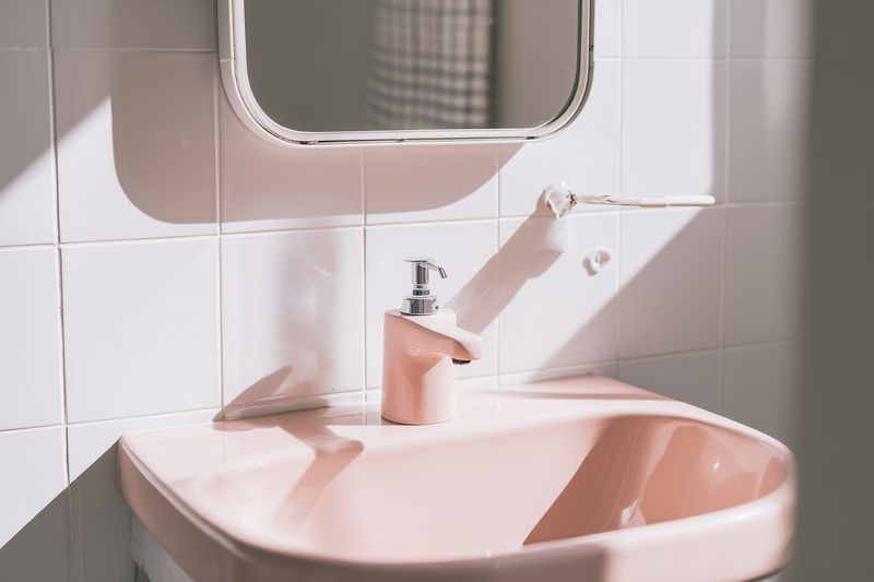 A sunlit pastel pink ceramic sink with chrome faucet against a white tiled wall, soft shadows and mirror reflection create a calm minimalist bathroom composition with gentle tones.