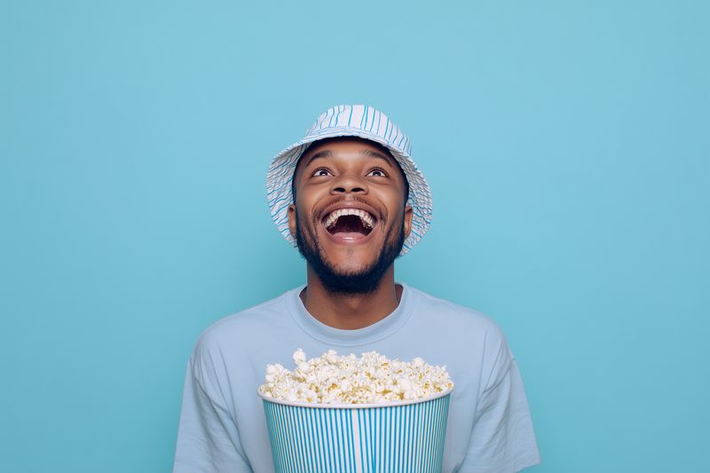 Young man wearing a striped bucket hat and casual t-shirt holding a large bucket of popcorn, laughing and looking upward against a bright blue studio background.