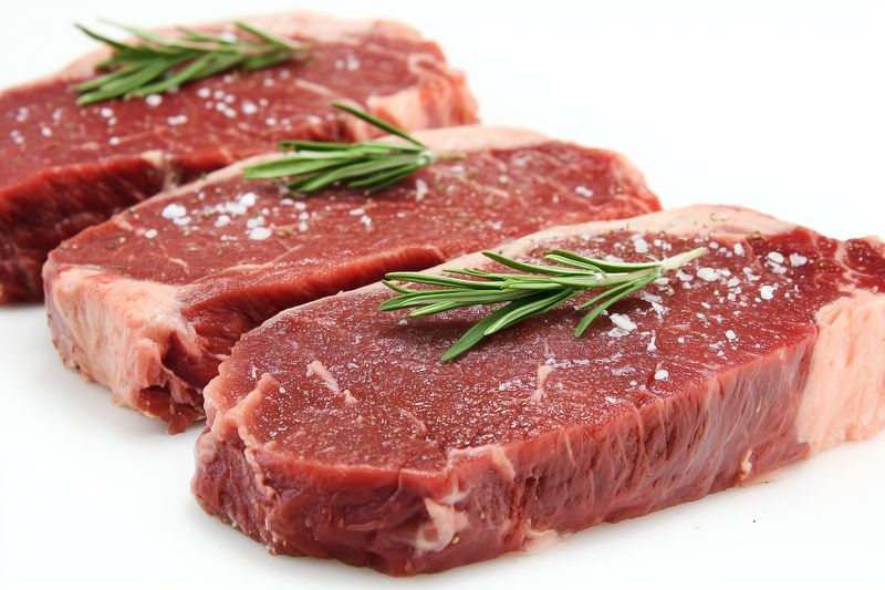 Three raw beef steaks seasoned with coarse salt and fresh rosemary sprigs arranged on a clean white background, closeup showing rich red color, marbling and texture for culinary use.