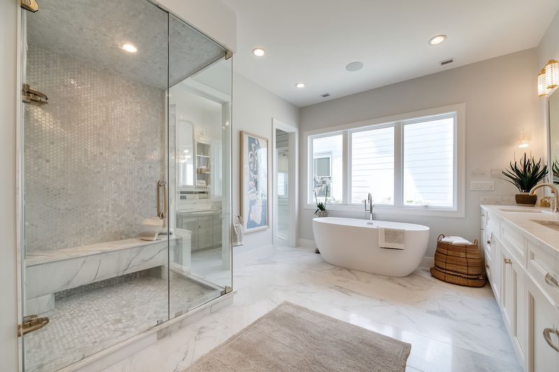 Bright modern bathroom interior featuring a freestanding soaking tub, glass shower enclosure, marble flooring and white vanity with large windows creating a serene, luxurious spa-like atmosphere.