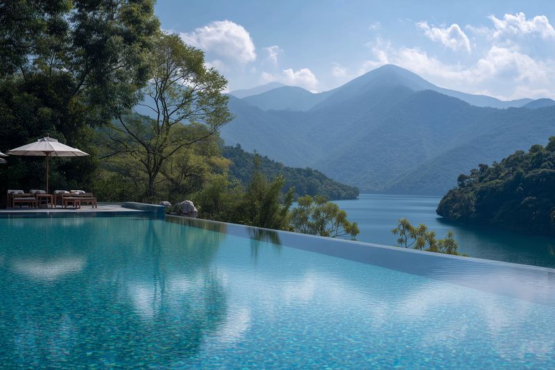 Infinity pool at a luxury resort overlooks a tranquil mountain lake surrounded by lush trees and distant peaks under a bright sky with soft clouds reflecting calm water.