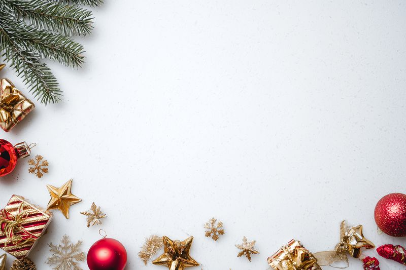 Festive holiday arrangement with red and gold Christmas ornaments, glittering stars, wrapped gift boxes and pine branches on a white textured background with ample negative space.