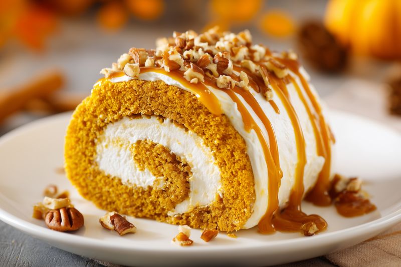 Closeup of a sliced pumpkin roll cake filled with creamy cheese frosting, drizzled with caramel and topped with chopped pecans on a white plate, autumn dessert vibe.